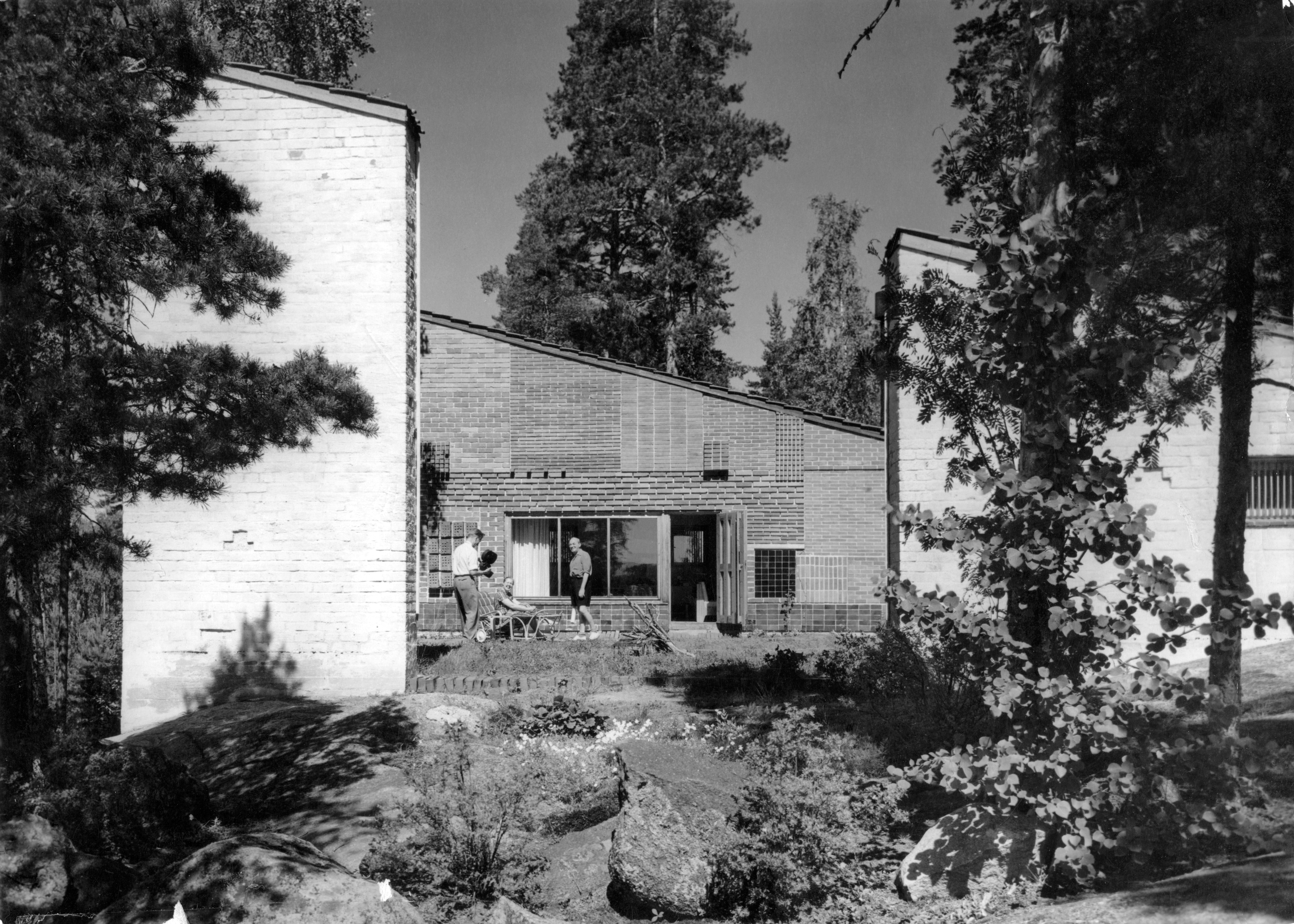 Mustavalkoinen ulkokuva Muuratsalon koetalosta. Alvar Aalto seisoo talon sisäpihalla.