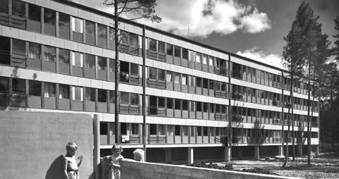 Eksteriöörikuva kerrostalosta. Etualalla kolme lasta ja taaempana rakennus mäntyjen ympäröimänä.