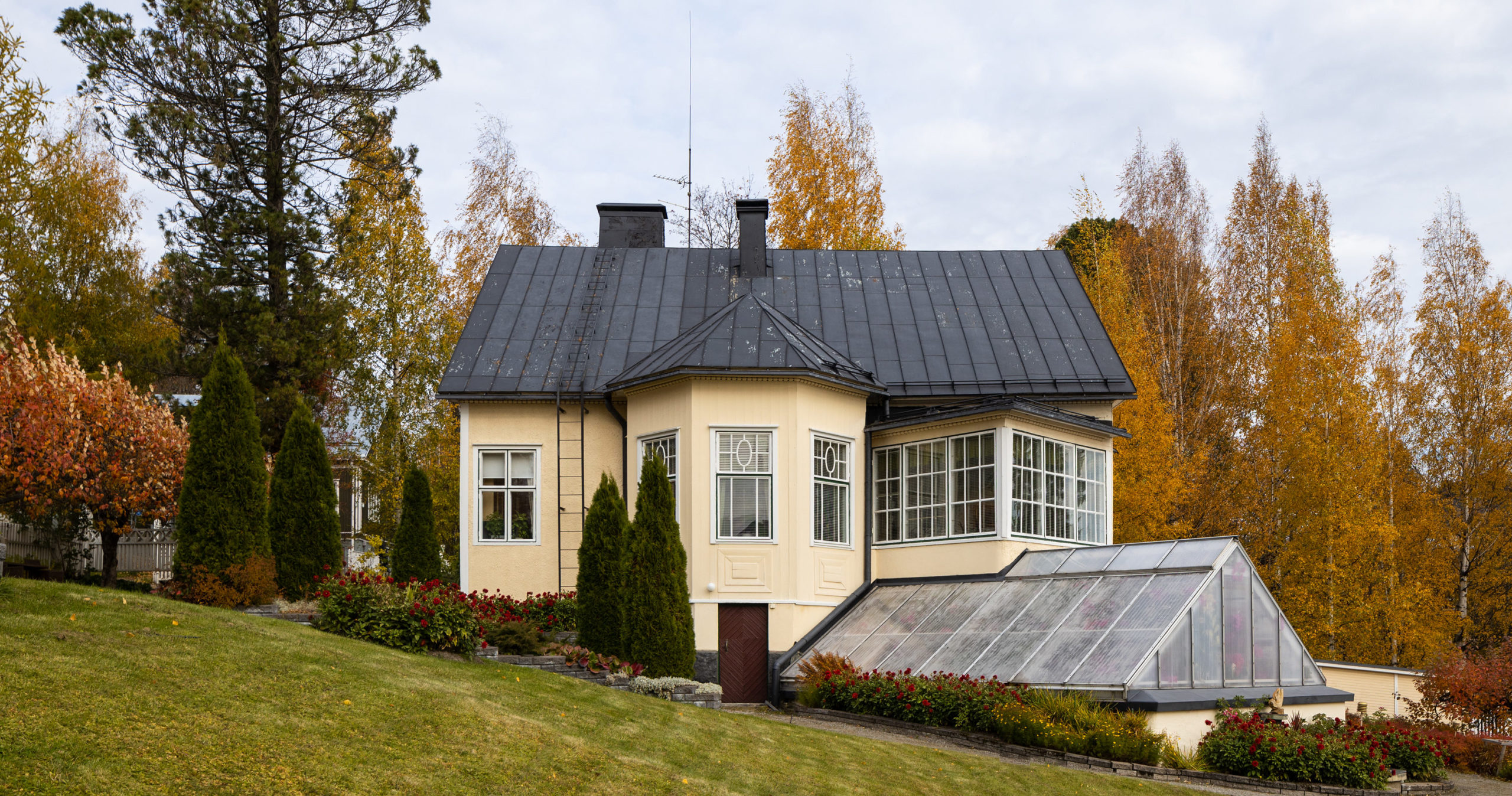 Keltainen rakennus syksyisessä maisemassa. Kuvan etualalla on hirveä nurmikko. Rakennuksen vieressä ja takana on erilaisia puita. Rakennuksen edustalla on kasvihuone.