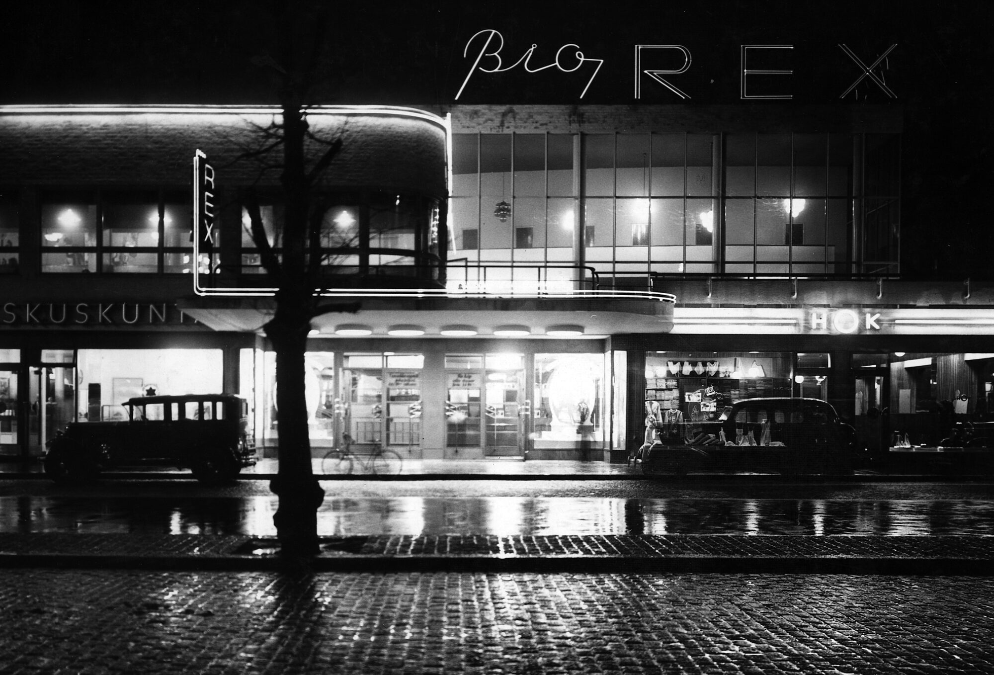 Matalan rakennuksen ikkunoista loistaa valoa pimeään ja sateiseen katukuvaan. Rakennuksen katolla on valomainos, jossa lukee "Bio Rex". Rakennuksen eteen on pysäköity muutamia autoja.
